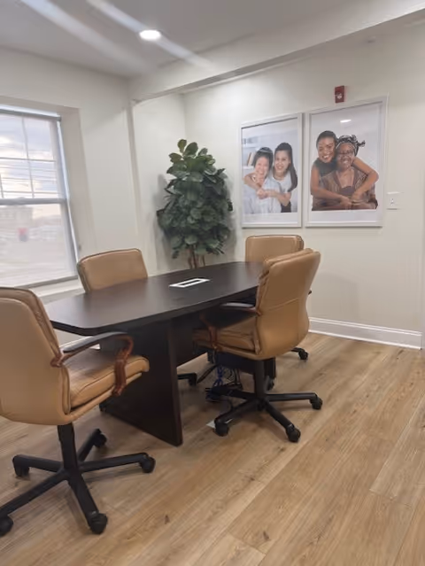 A conference-style room with a dark table, tan chairs, potted plant, and wall photos.