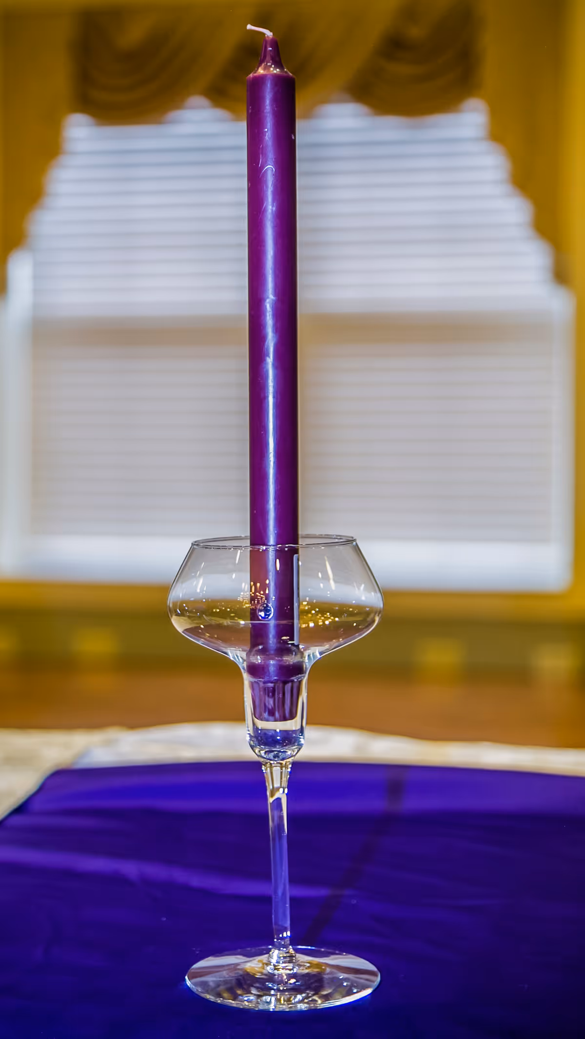 A tall purple candle in a glass holder on a table with a purple cloth.