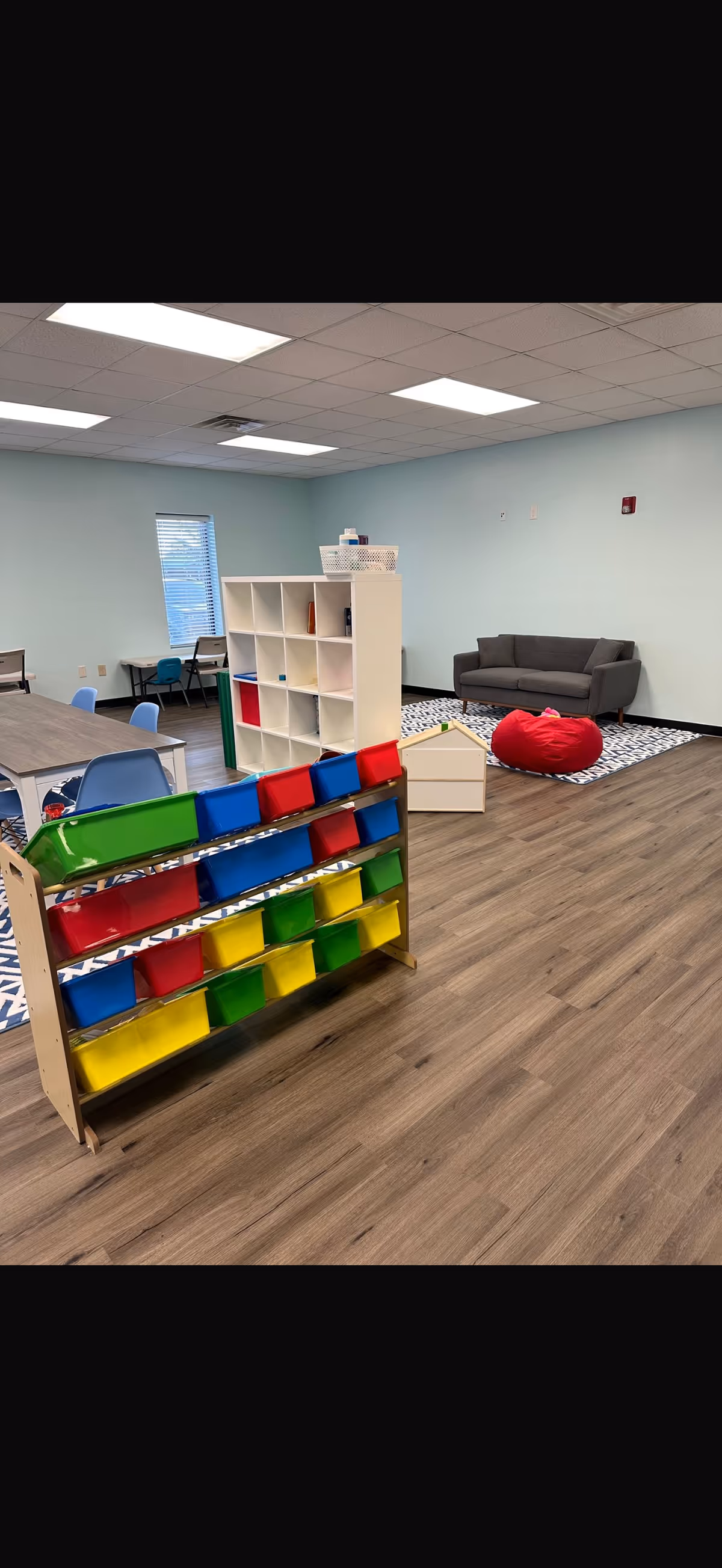 Bright interior playroom with colorful bins, shelves, tables, and a gray couch.