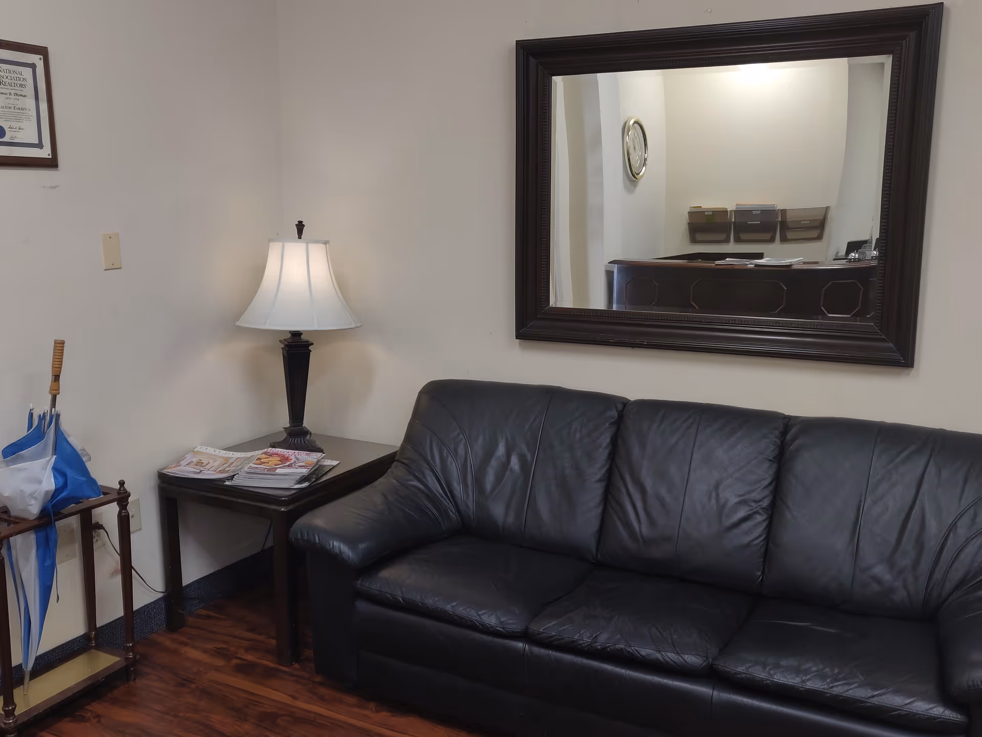 A black leather sofa with a side table, lamp, and wall mirror in a waiting area.