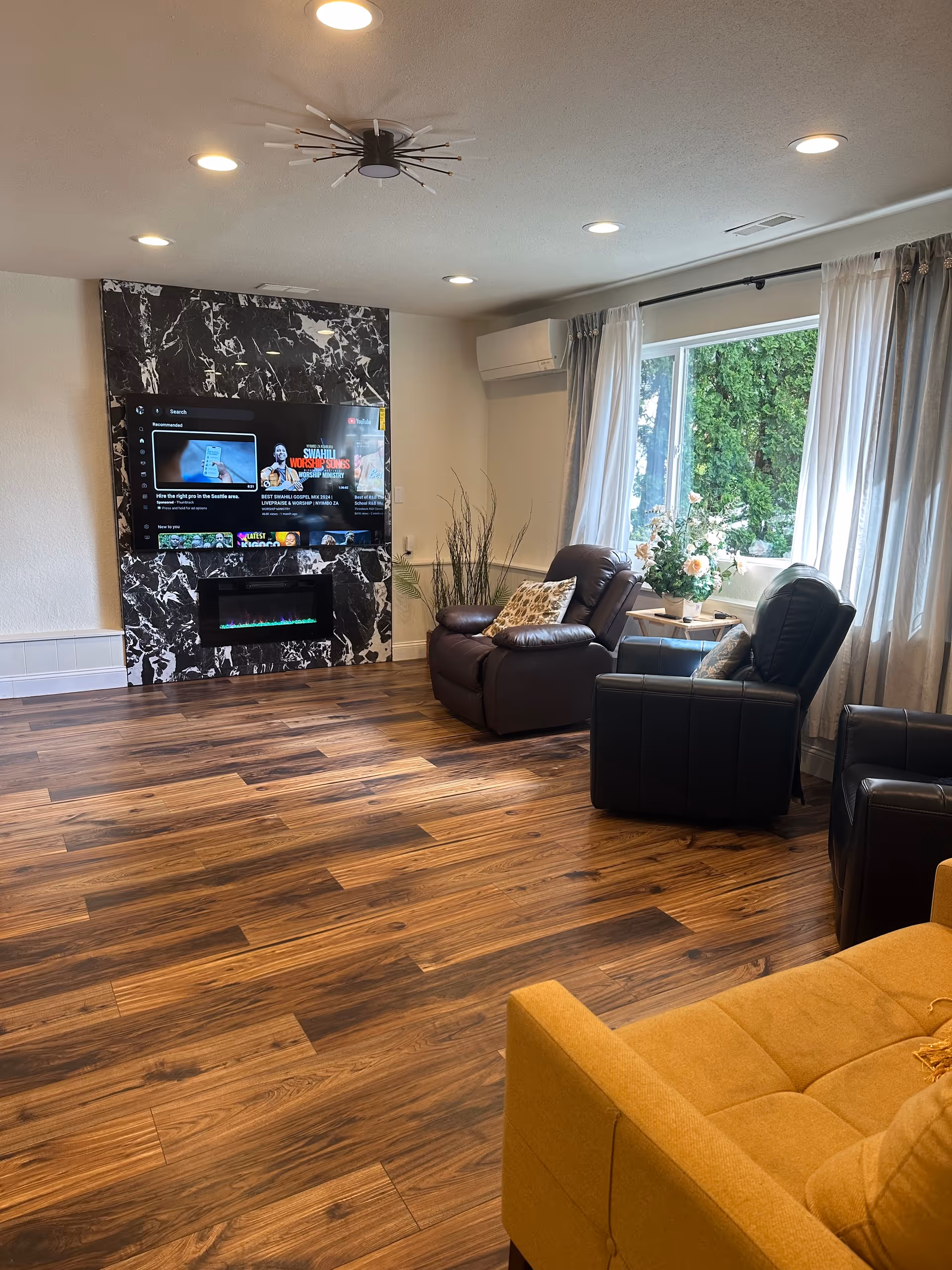 Spacious living room with a large TV on marble panel, leather recliners, and sunny windows.