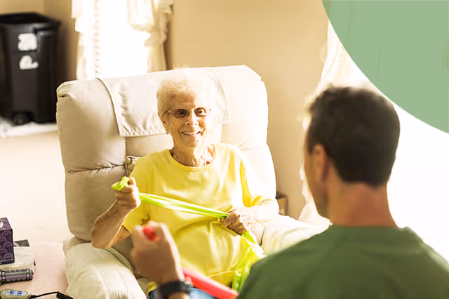 Smiling elderly woman in a recliner chats with a caregiver in a living room.
