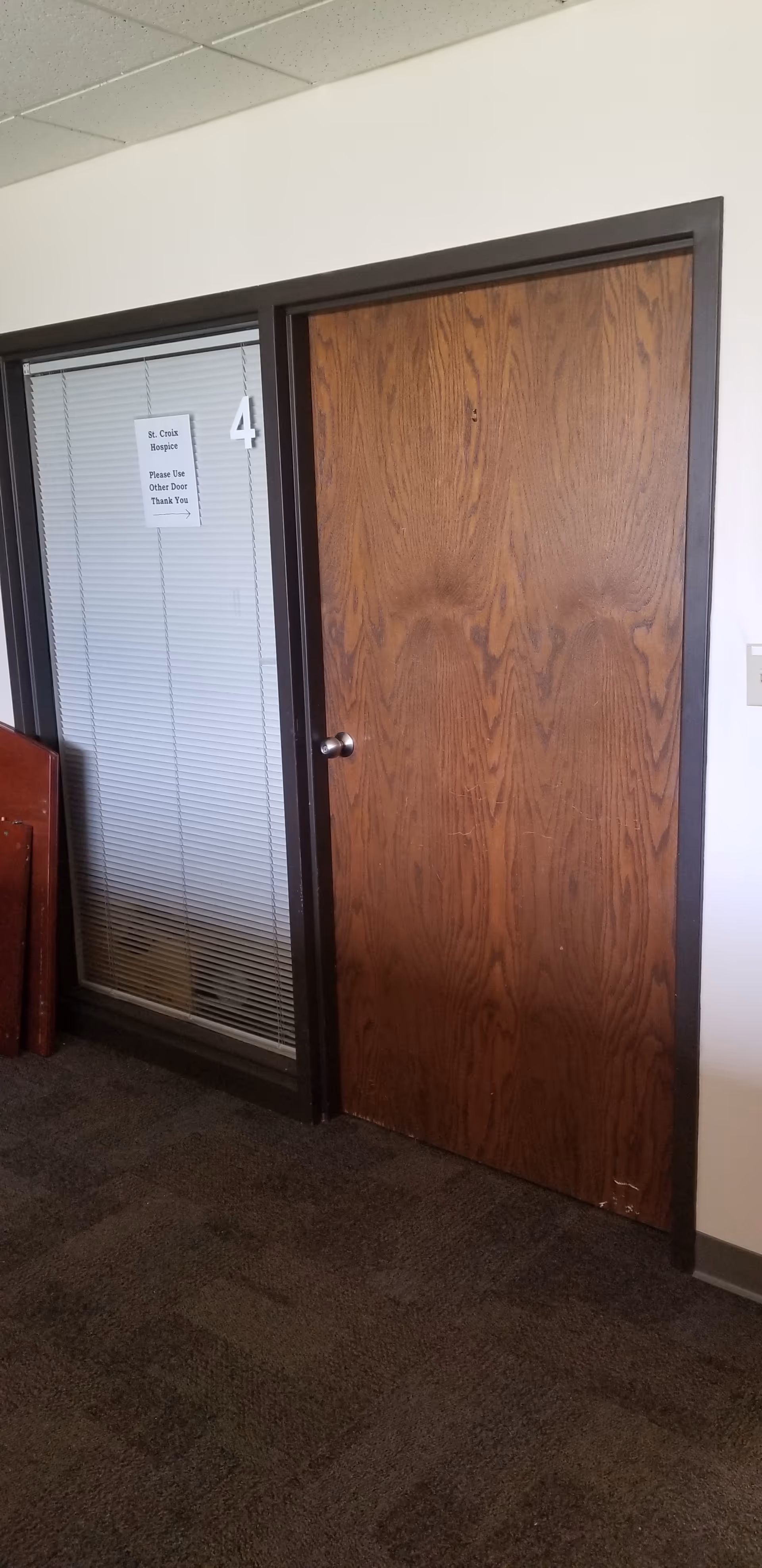 Wooden door and glass doorway with blinds in a carpeted hallway of a senior care facility.