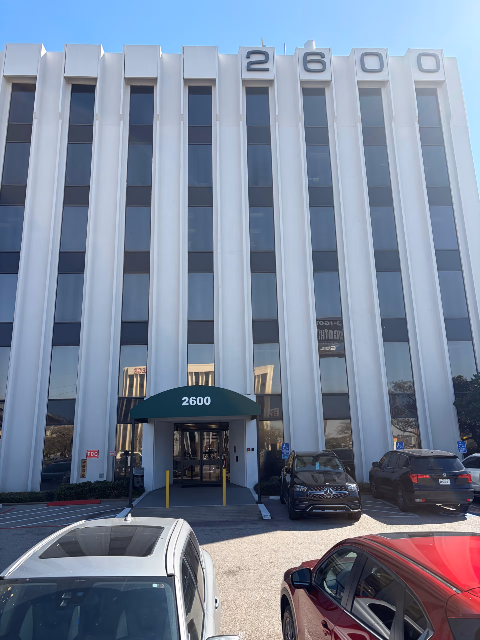 Front of a tall white building with a green entrance canopy and parked cars.