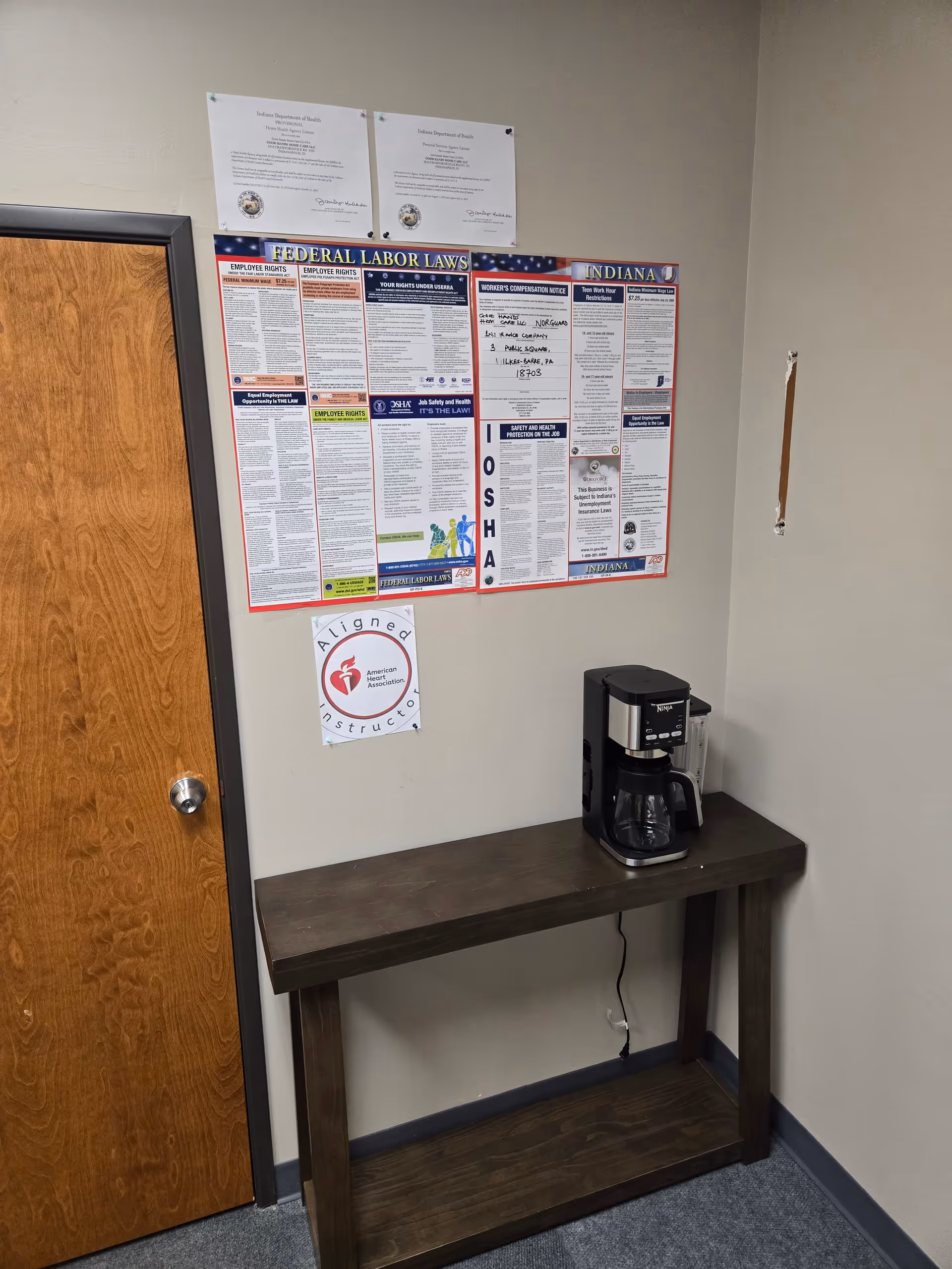 Interior corner with wooden table, coffee maker, and safety/poster boards on the wall.