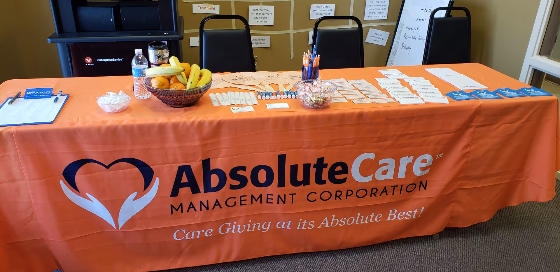 Table covered with orange AbsoluteCare banner, informational materials, fruit basket, and chairs in a lobby.