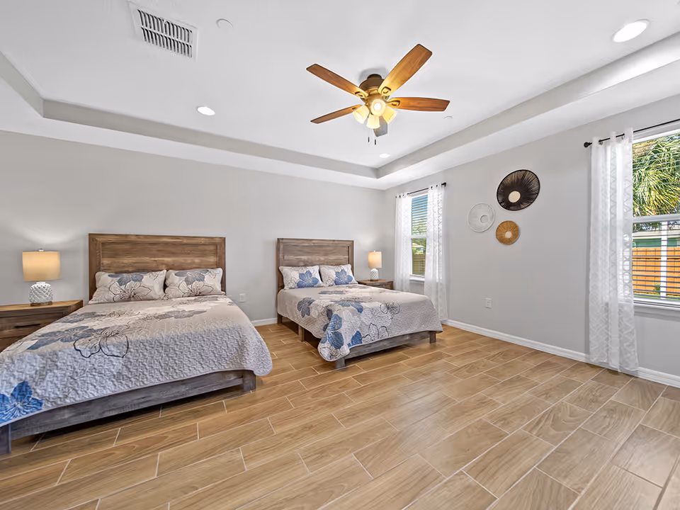 Two beds with matching nightstands, a ceiling fan, and windows in a bright bedroom.