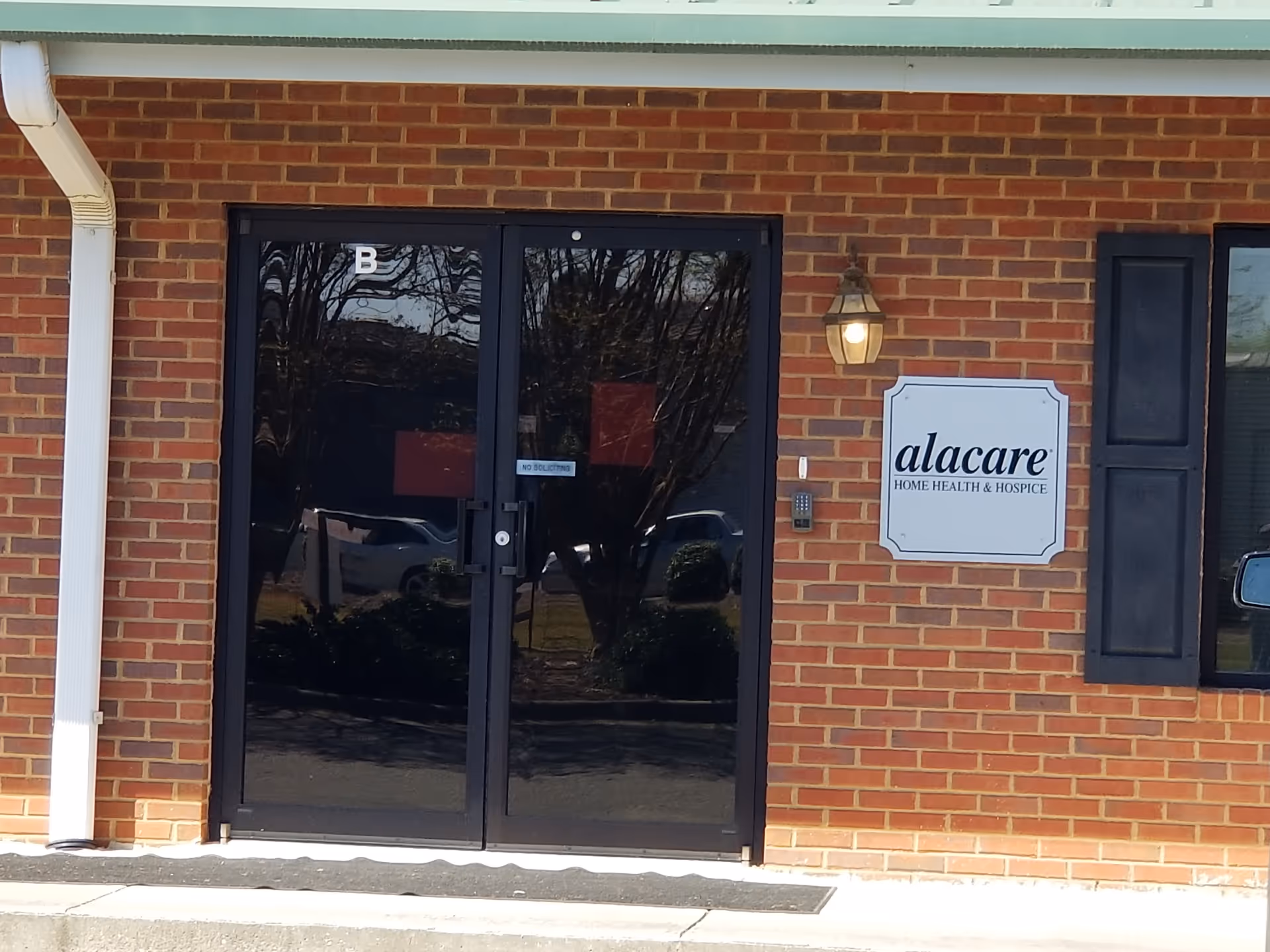 Brick building exterior with glass doors and alacare sign beside a wall light.