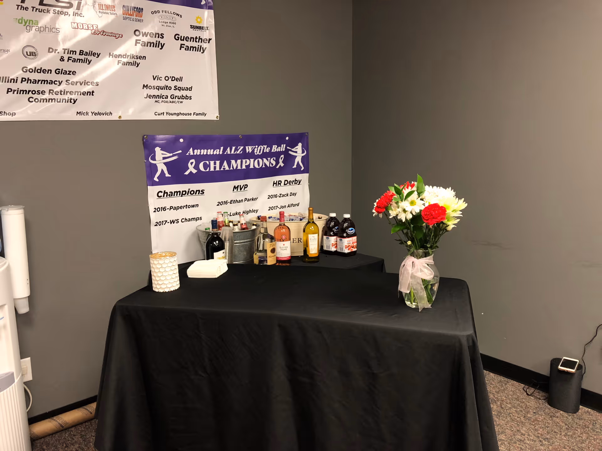 Indoor event setup with a black tablecloth, banners, beverages, and a vase of flowers.