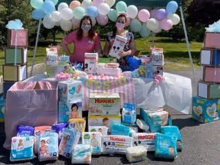 Two masked caregivers stand behind a table of diapers and gifts at an outdoor donation event.
