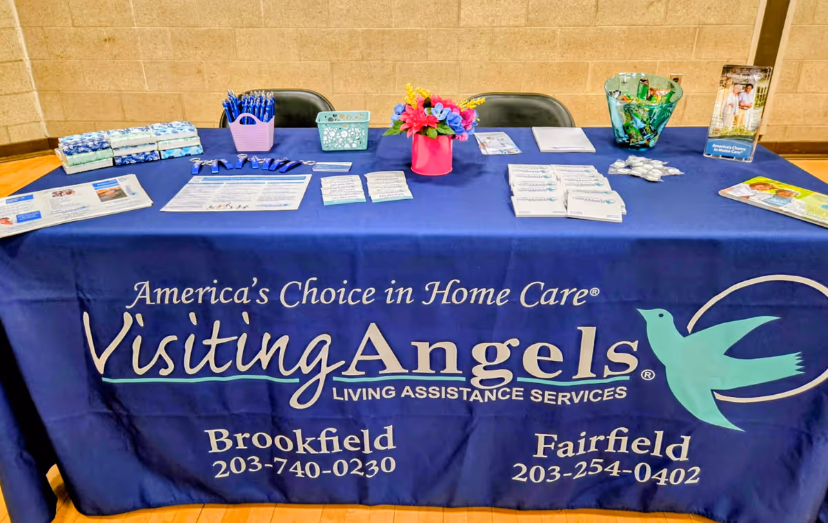 Promotional table indoors with a blue Visiting Angels banner and assorted brochures and items.