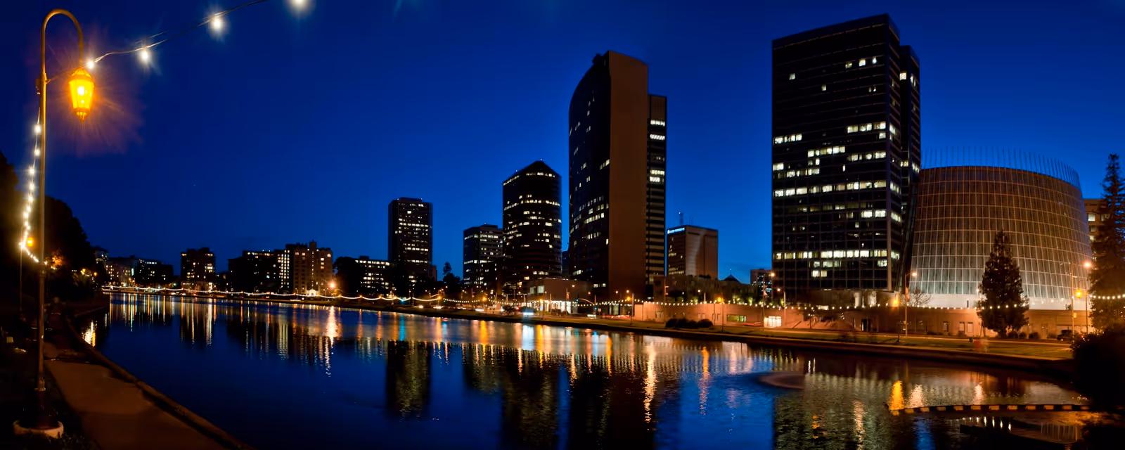 Nighttime city skyline reflects on calm river with illuminated buildings and string lights.