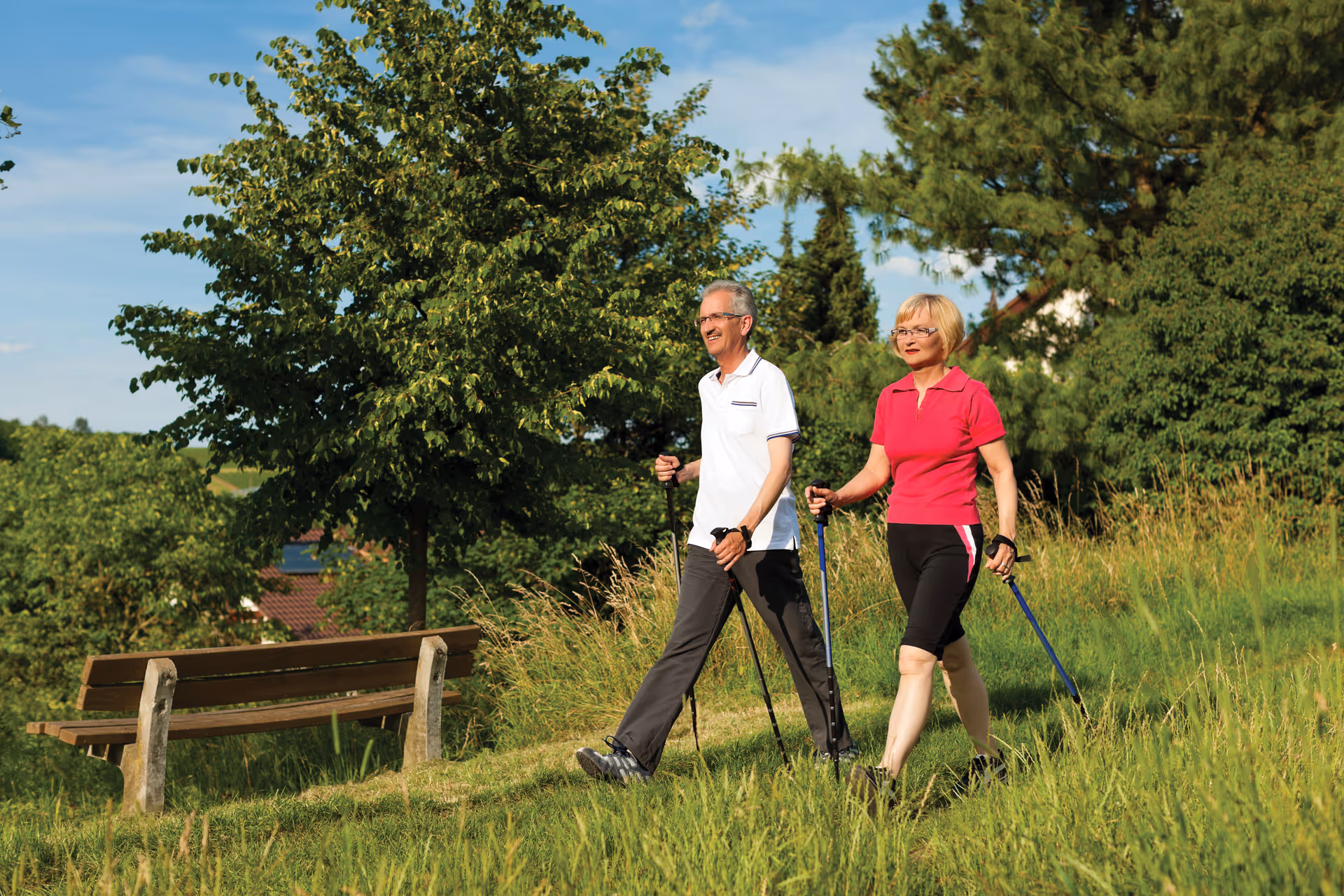 Two seniors walk with trekking poles on a sunny grassy outdoor area beside a bench.