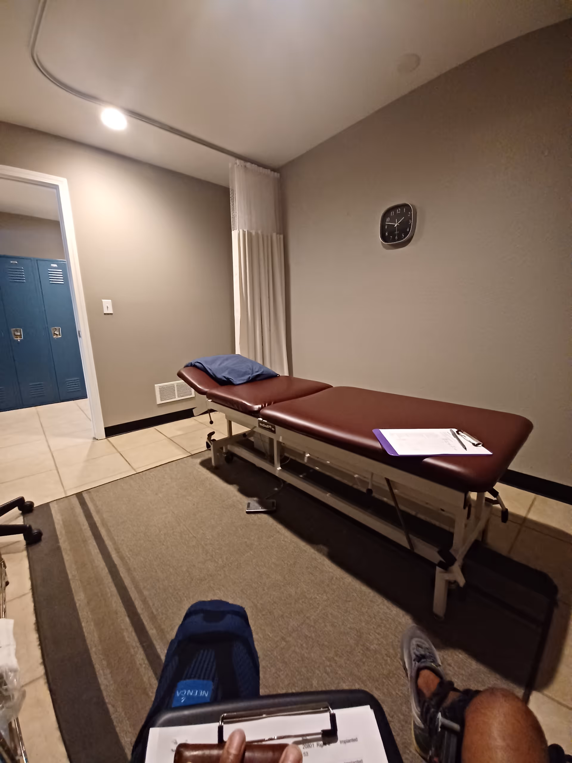 Treatment table in a small clinic room with clock, curtain, and blue lockers visible.