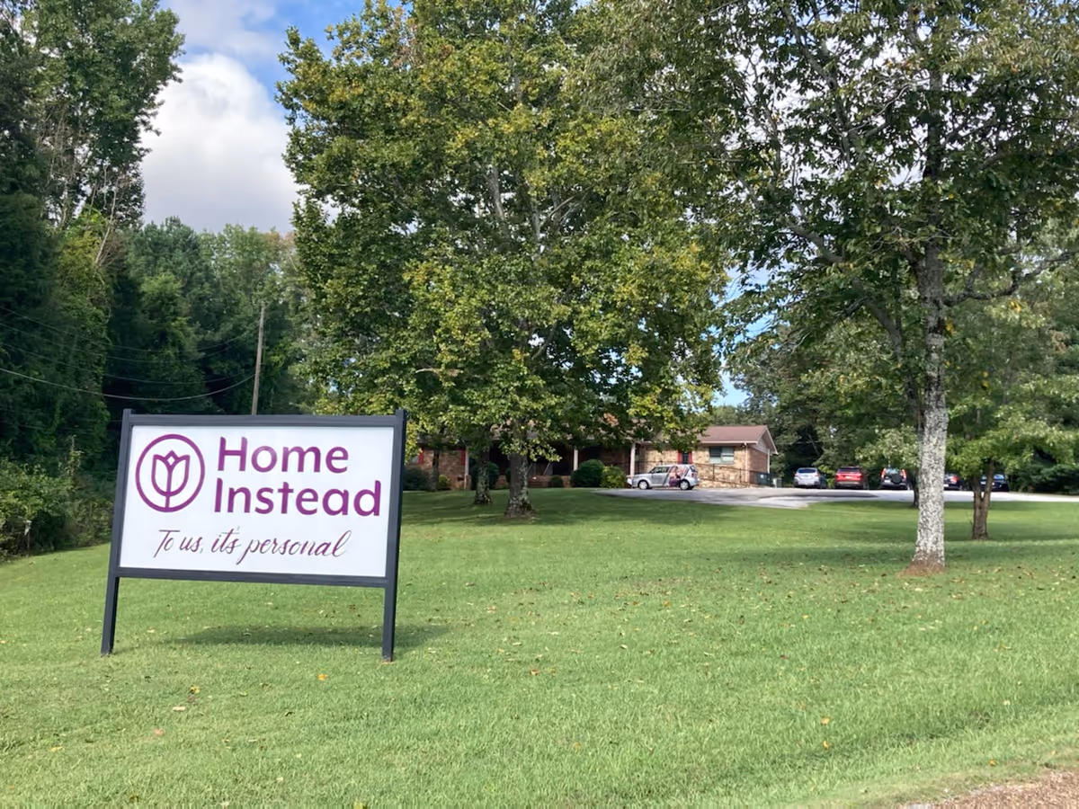 A Home Instead sign sits on a grassy lawn with a brick building behind it.