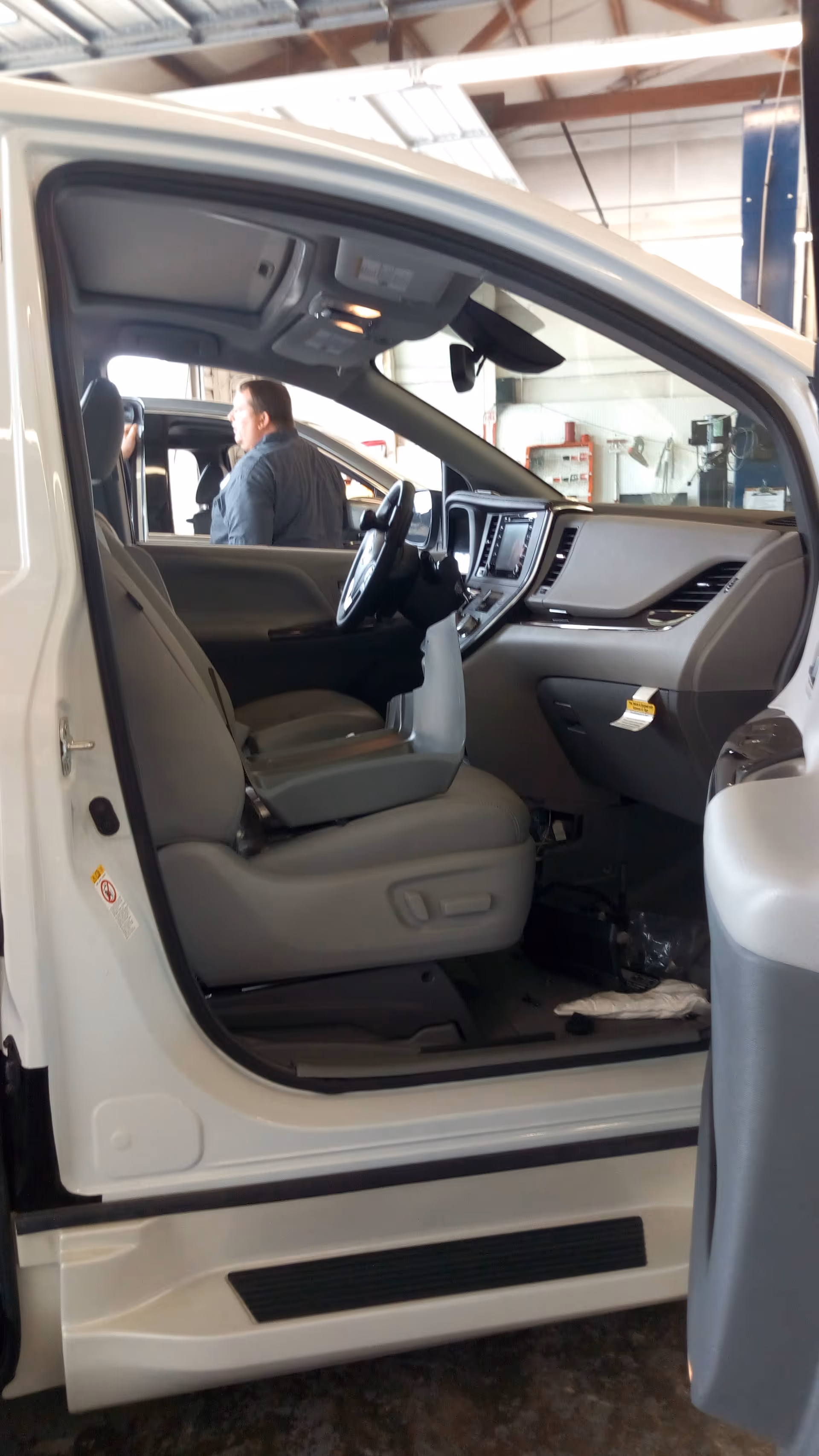 Interior of a white van showing driver seat, dashboard, and a mechanic in the background.