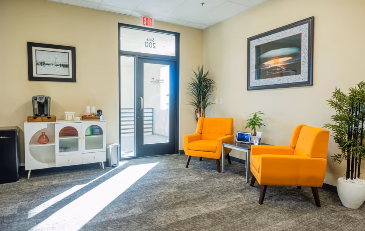 Bright lobby seating area with orange chairs, plants, coffee station, and exit door.