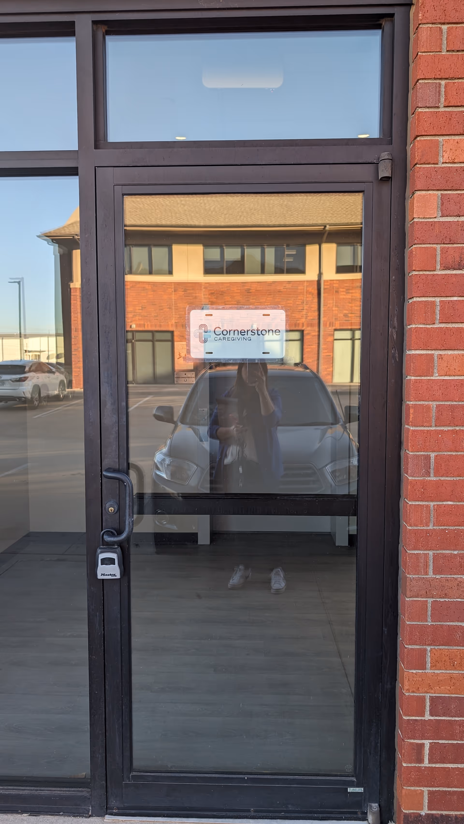 Glass entrance door shows Cornerstone Caregiving sign; reflection of photographer, brick exterior.