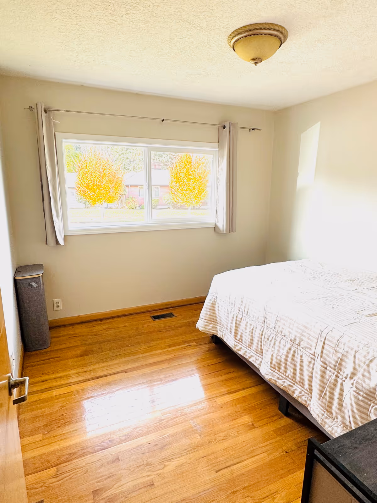 A small bedroom with a bed, hardwood floor, window with curtains, and minimal furniture.