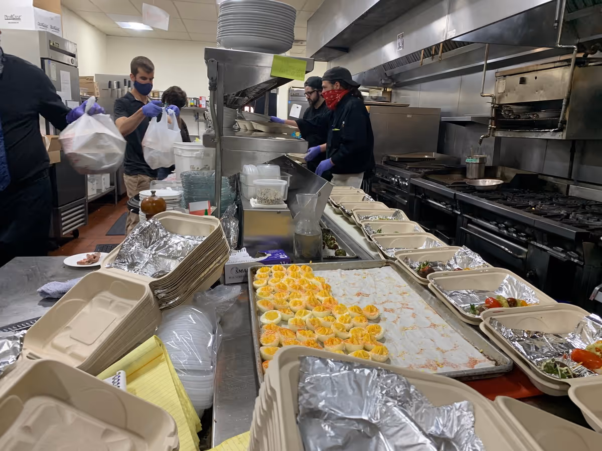 Kitchen scene with staff packing meals, trays of food, and stainless appliances.