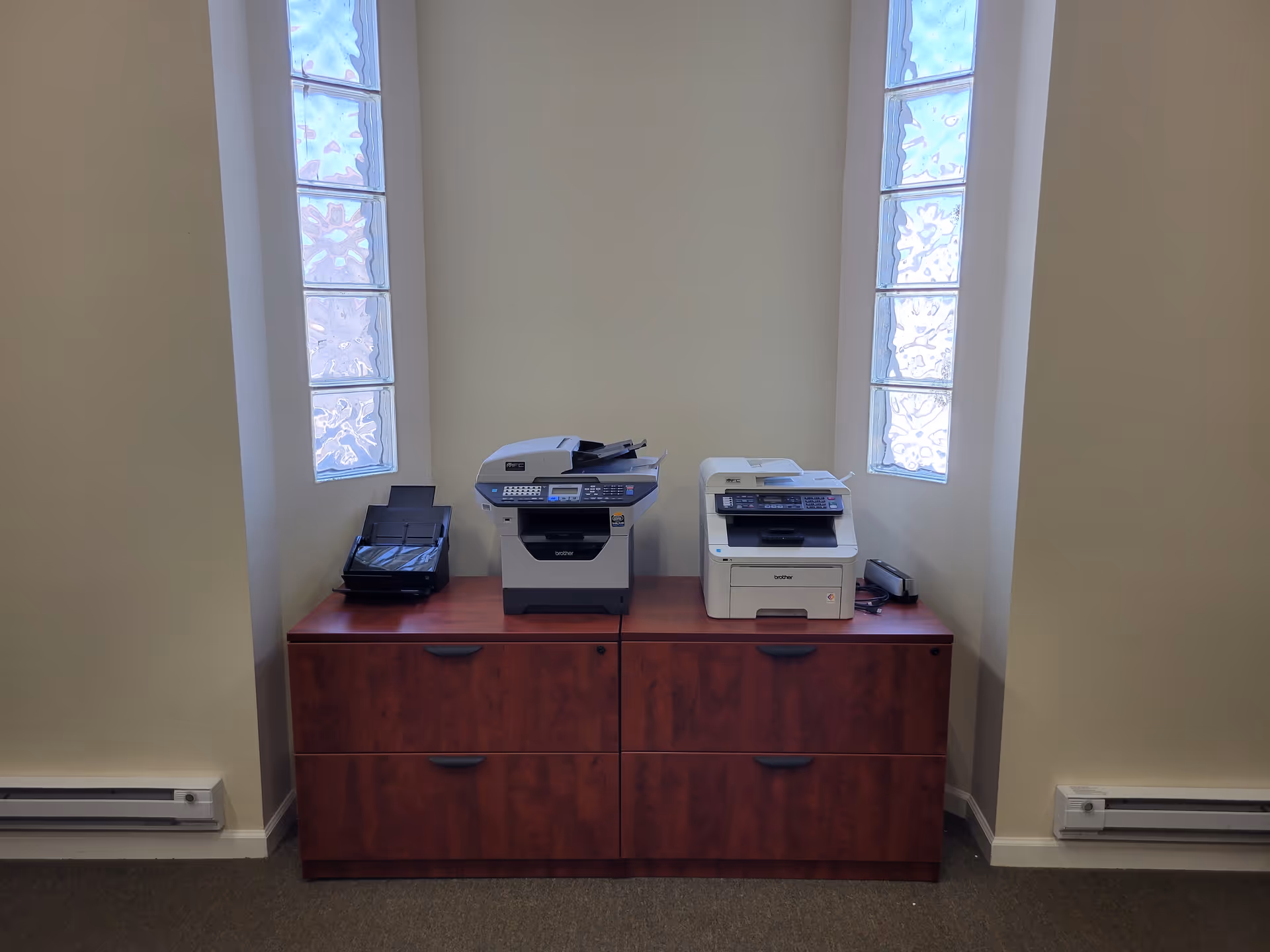 Two printers sit on a wooden cabinet in a narrow interior corner by glass block windows.