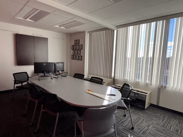 A conference room with a long oval table, several chairs, two computer monitors, and large windows.