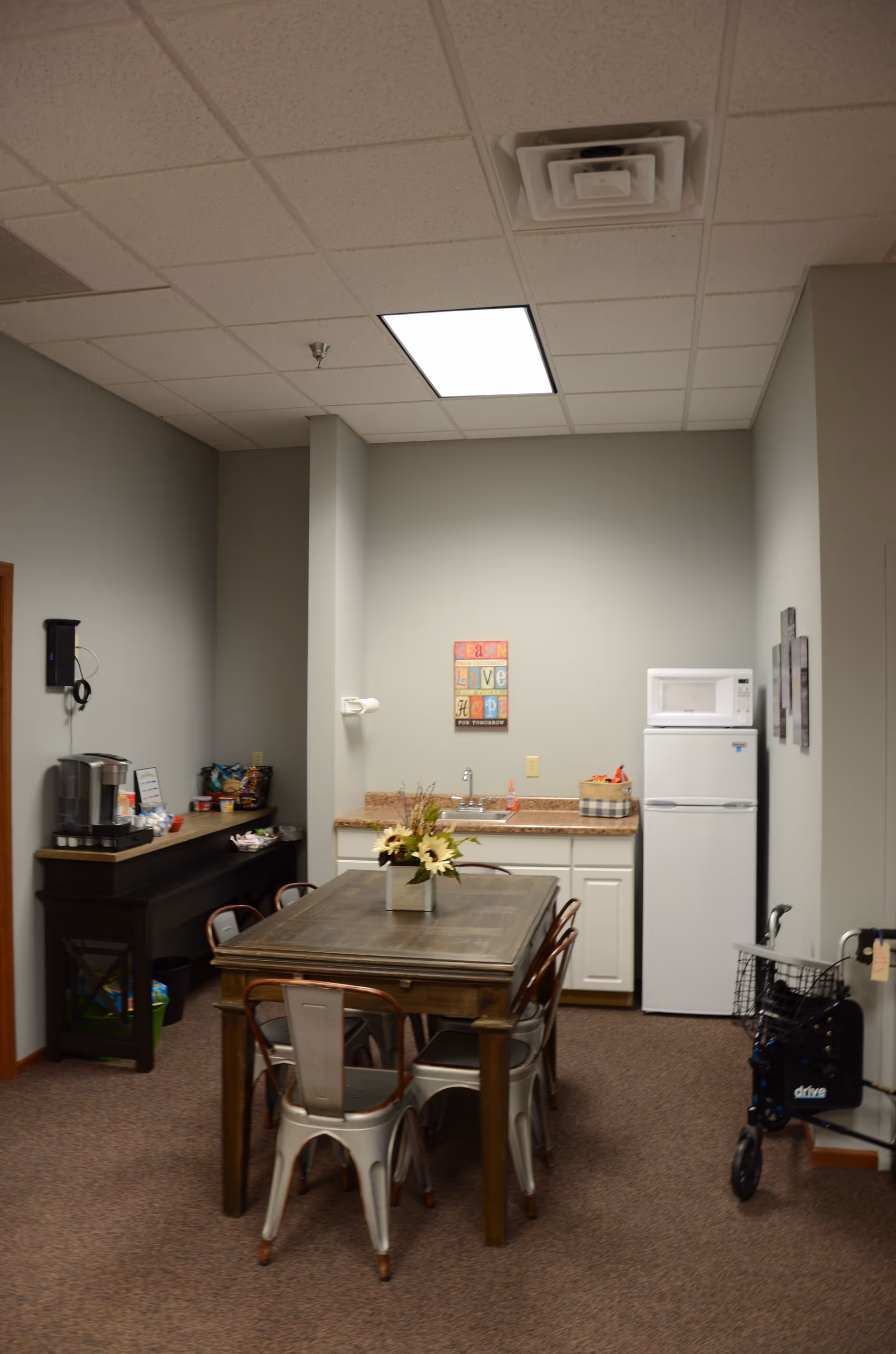 Communal dining area with a wooden table, metal chairs, kitchenette, and a walker nearby.