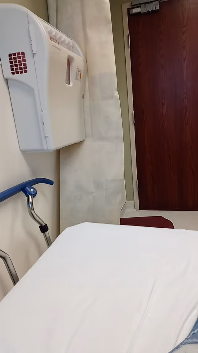 Interior patient room with a bed, wall-mounted medical device, blue walker, and wooden door.