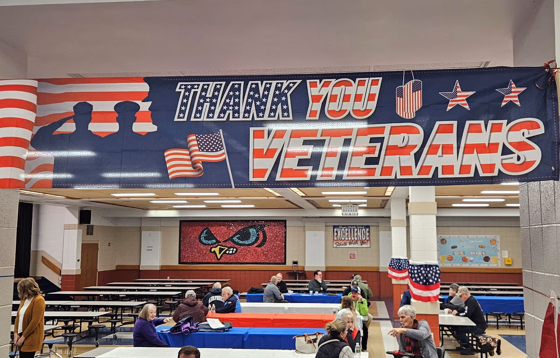 Indoor dining hall with a large 'Thank You Veterans' banner and attendees seated at tables.