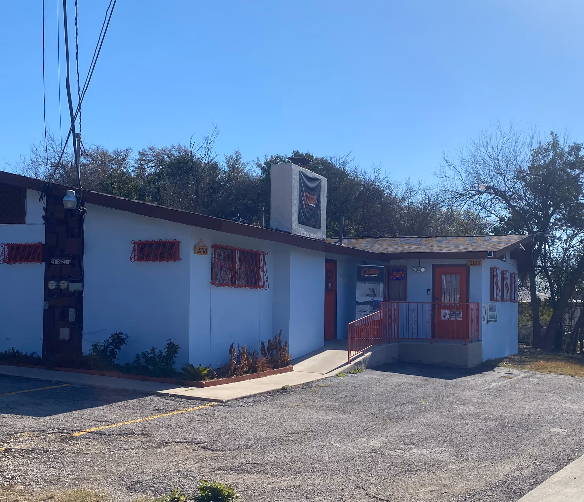 Small light-blue building with red doors and railings, parking lot in the foreground.