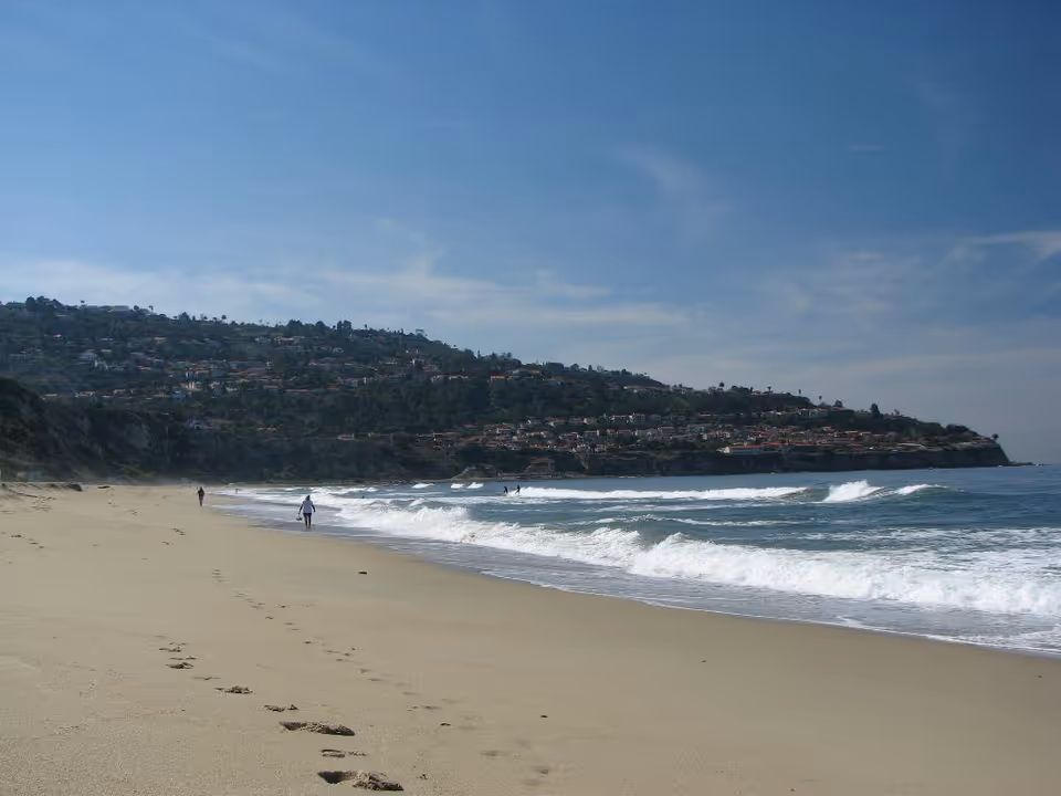 Sunny beach with footprints, waves crashing, and a hillside town in the distance.