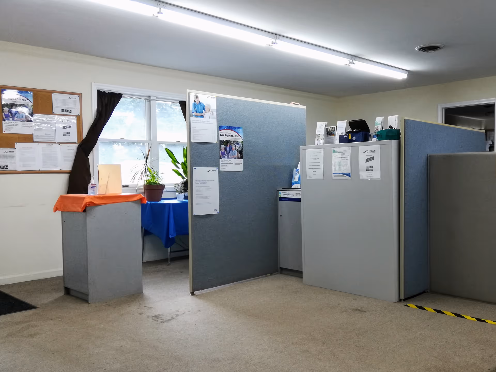Interior of a LINCARE facility with partitioned cubicles, bulletin boards, and potted plants.