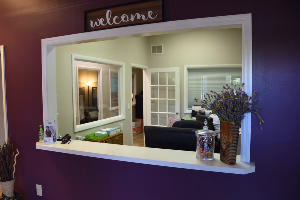 Purple-walled reception window with counter, a welcome sign, and a lavender-filled vase.