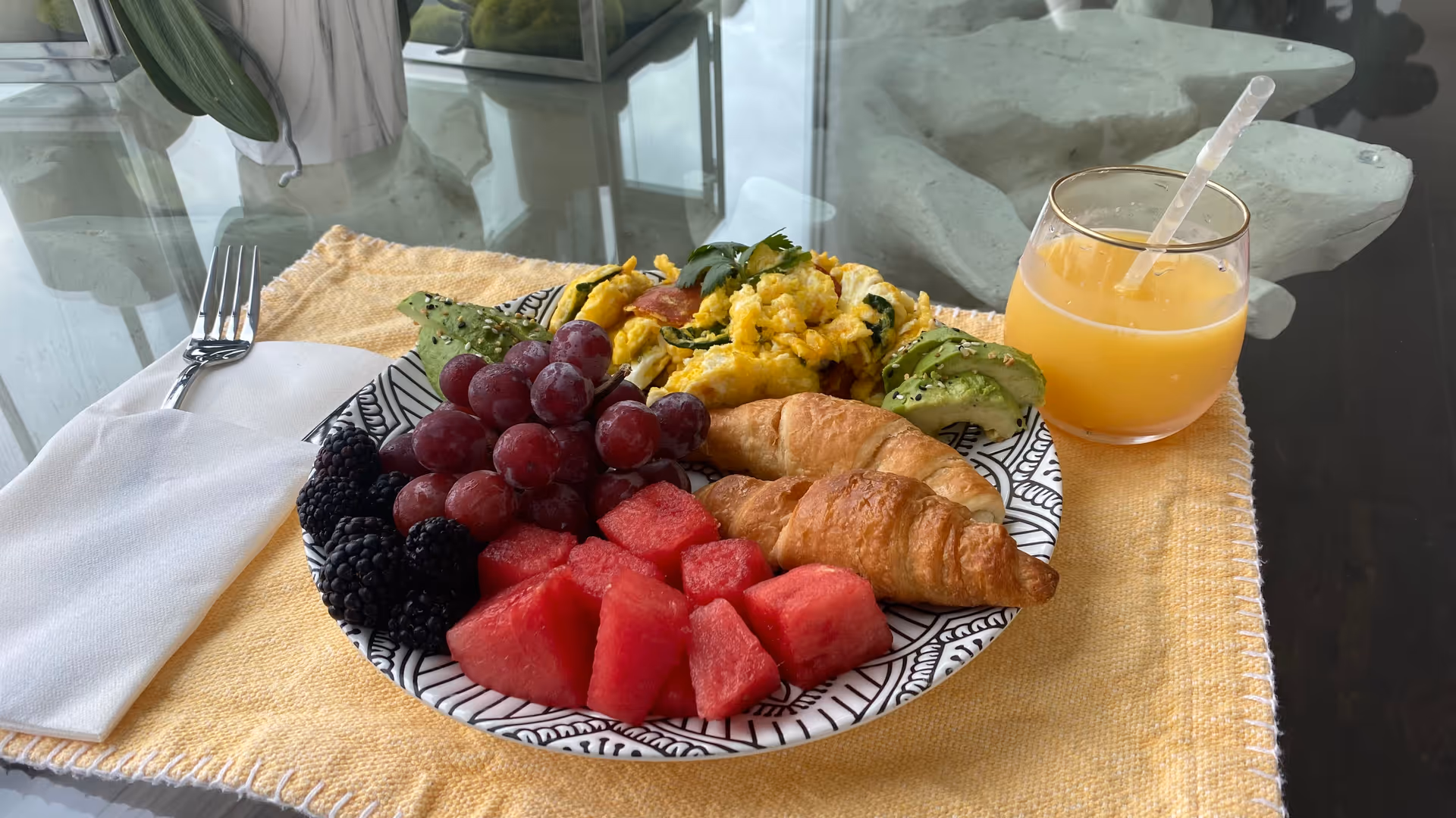 Plate of fruit, croissant, scrambled eggs, and juice on a table by a window.