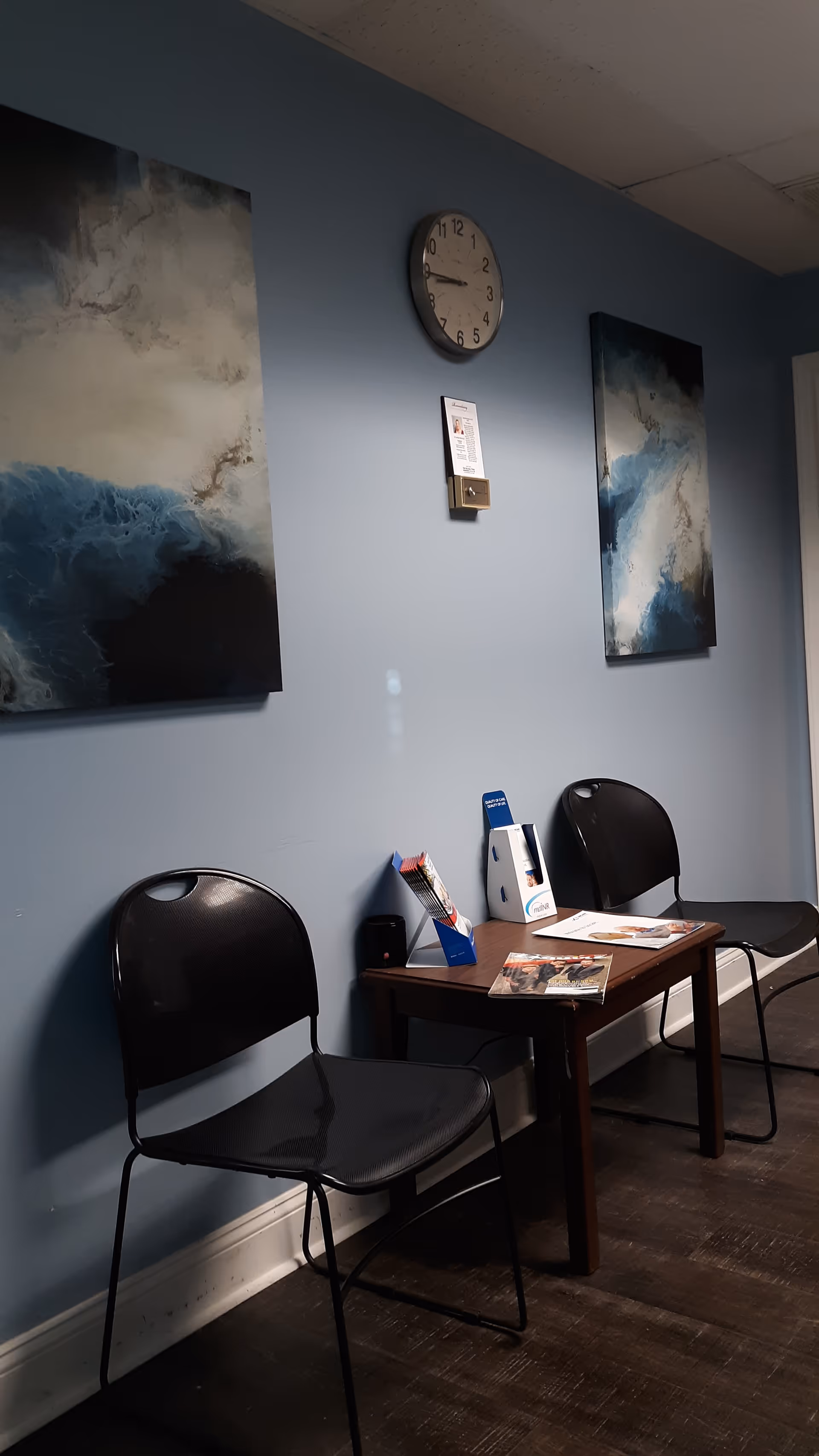 Blue-walled waiting area with two black chairs, a small table, brochures, a clock, and wave artworks.
