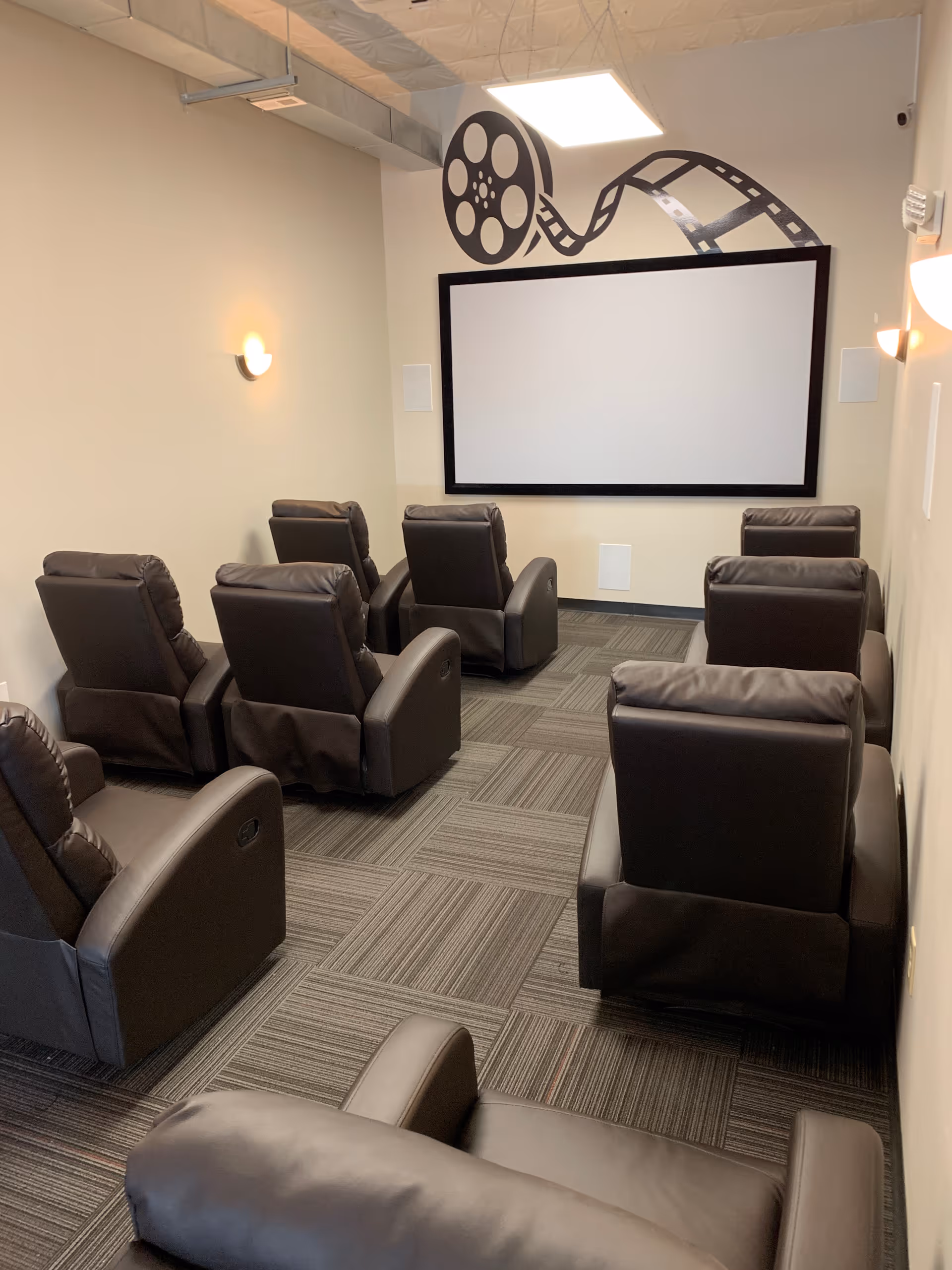 Small theater room with black leather recliners facing a screen and film reel mural.