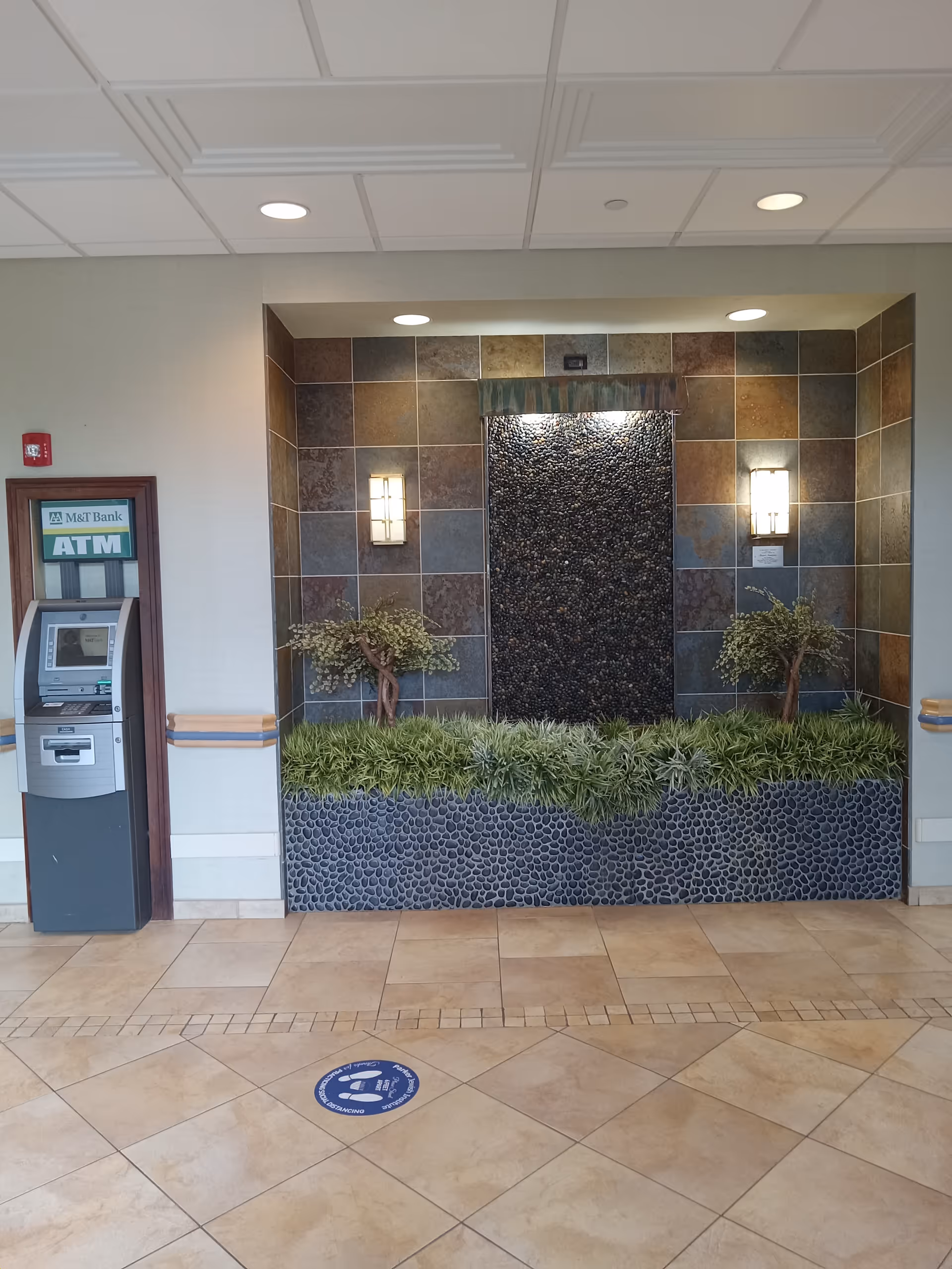 Lobby interior with pebble fountain, tiled wall, greenery, and an ATM on the left.