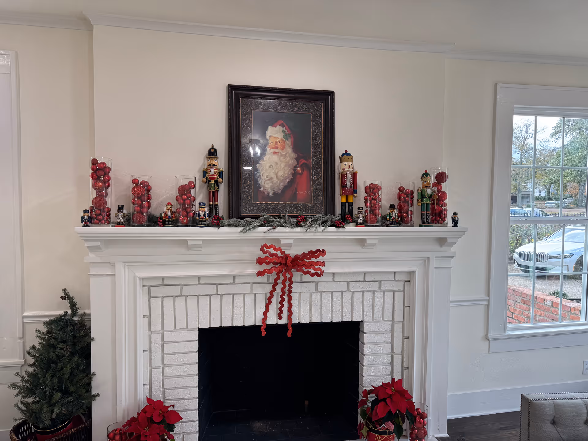 White fireplace mantel adorned with red ornaments, garland, nutcrackers, and Santa painting.