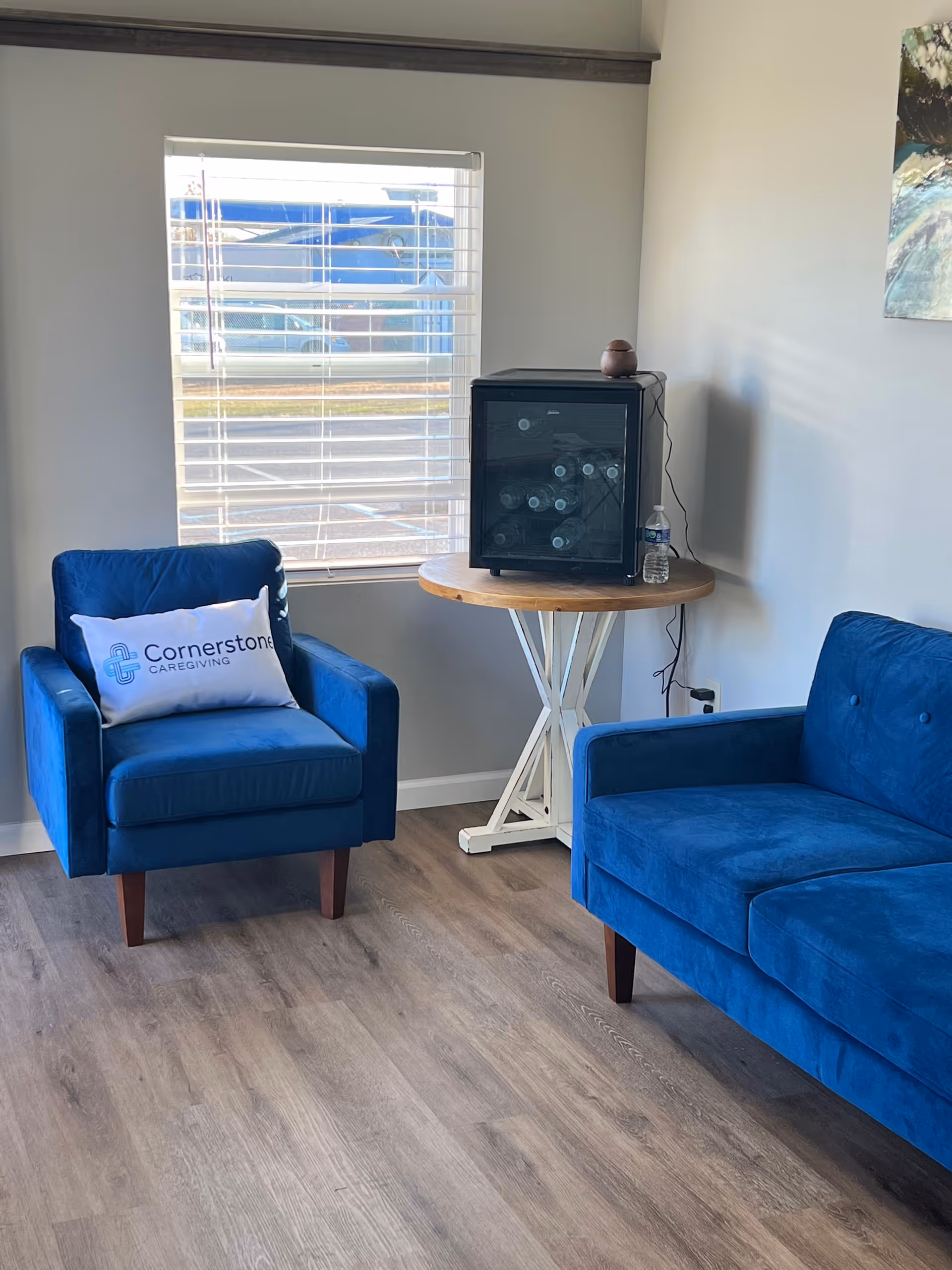 Bright living room with blue chairs, a round table, mini fridge, and Cornerstone pillow.
