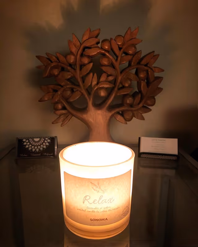 A glowing candle sits on a glass table with a wooden tree sculpture behind.