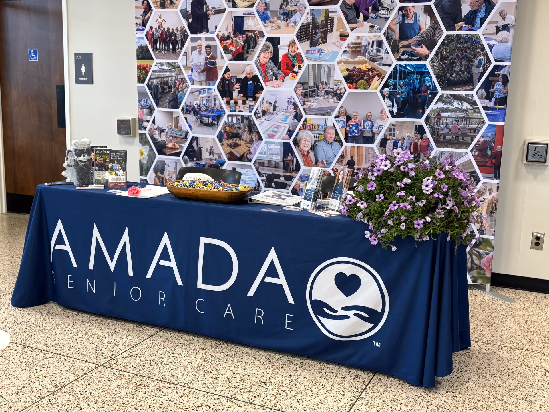 A blue Amada Senior Care table in a lobby with a honeycomb collage backdrop and flowers.
