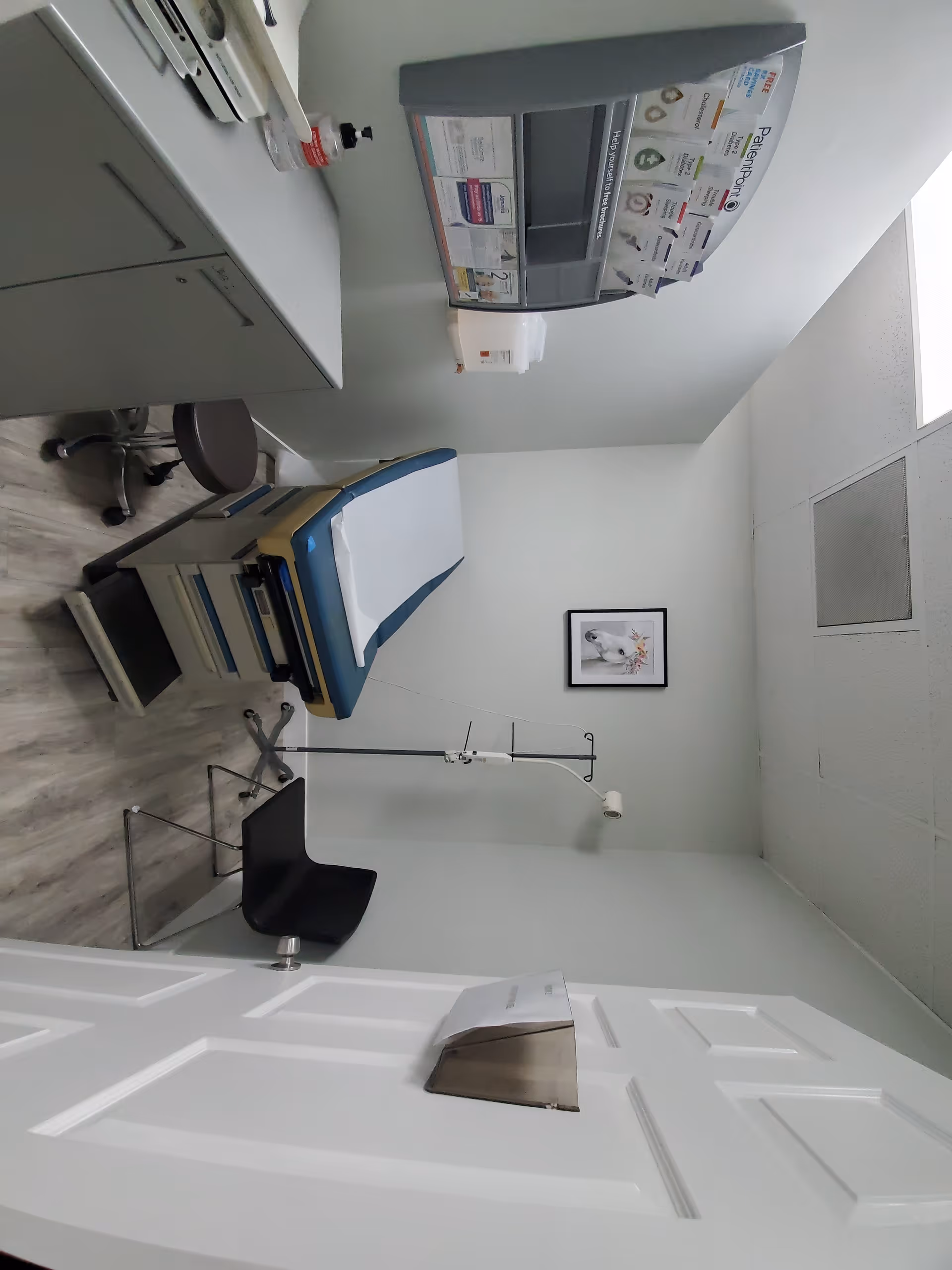 A clinic examination room featuring a hospital bed, medical equipment, cabinets, and wall art.