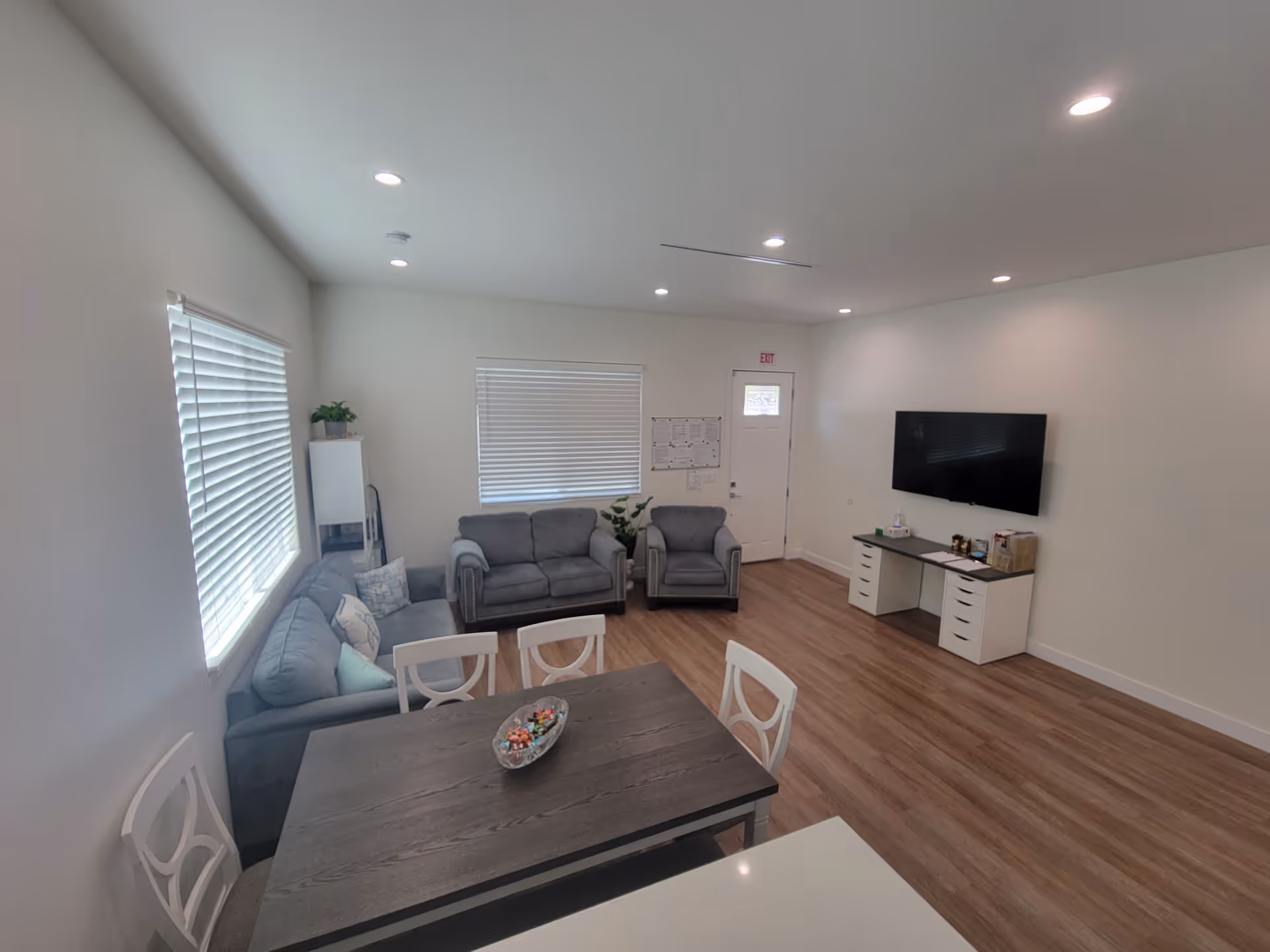 Bright, modern senior living common area with dining table, couches, and a wall-mounted TV.