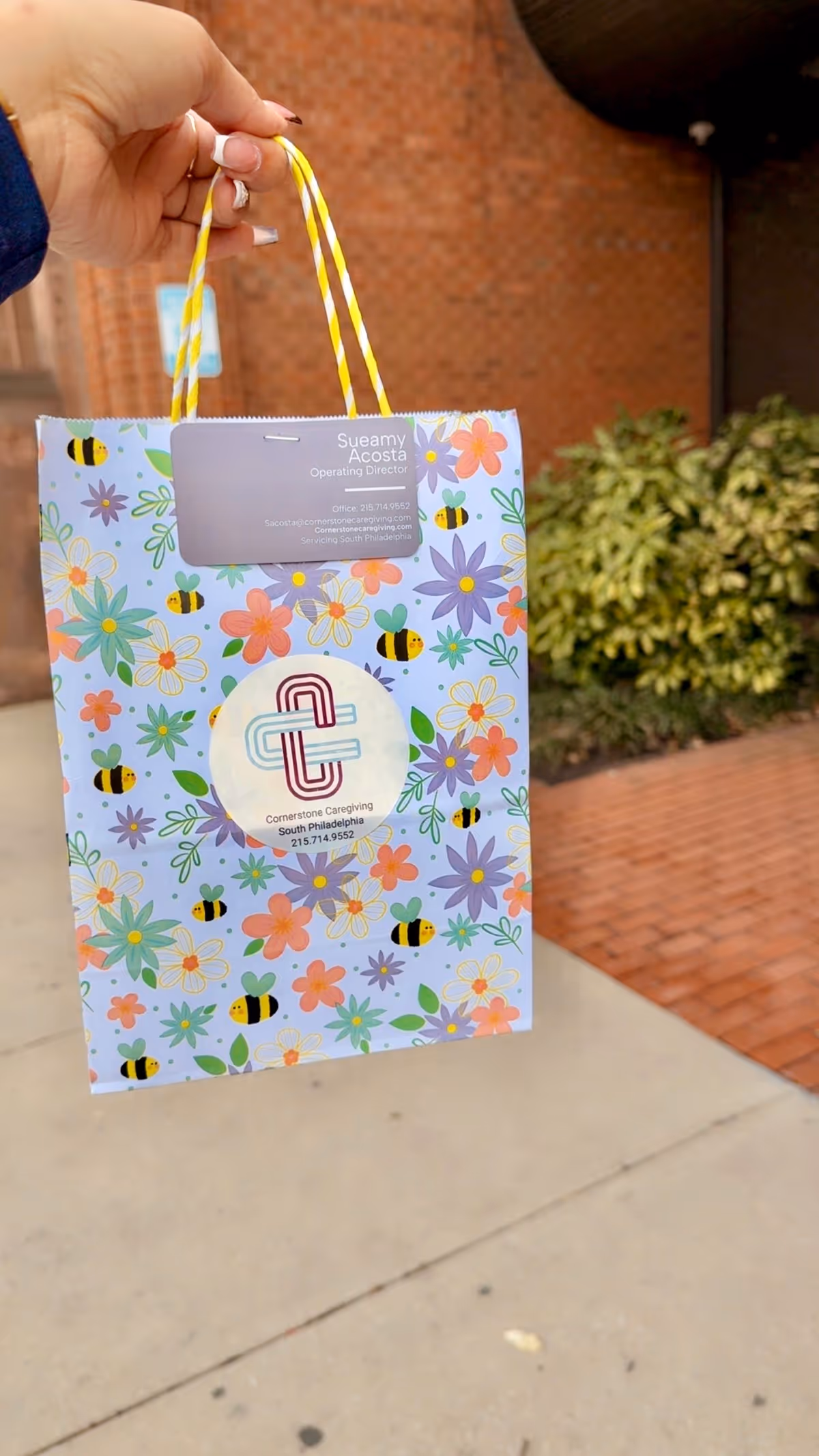 A hand holds a colorful floral gift bag outside Cornerstone Caregiving facility.