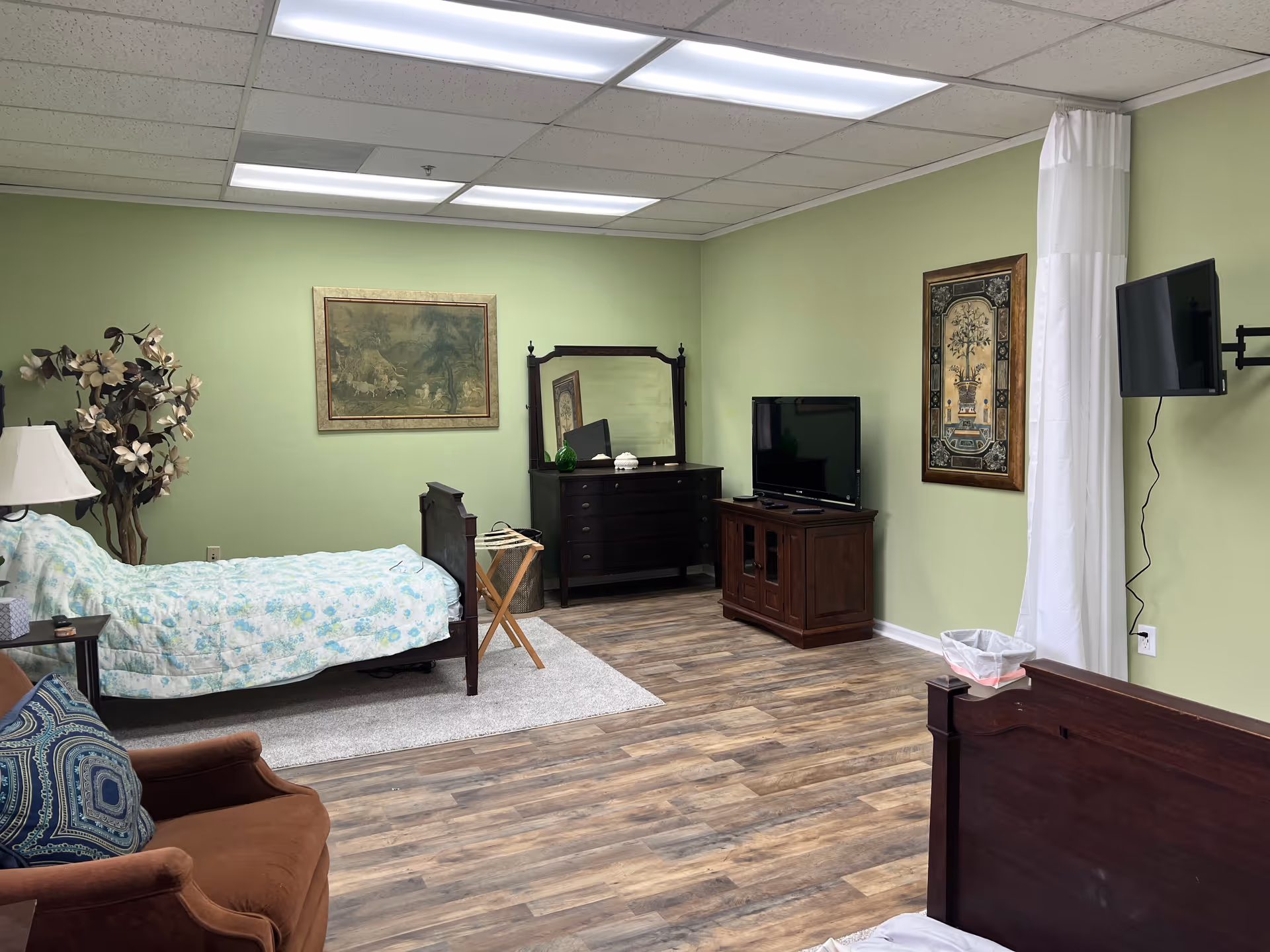 A furnished senior living bedroom with a bed, dresser, television, armchair, and green walls.
