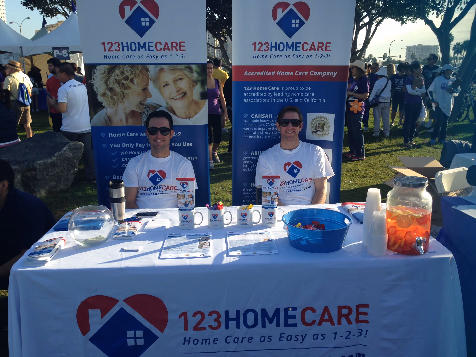 Two staff members sit at a table with 123 Home Care banners outdoors.