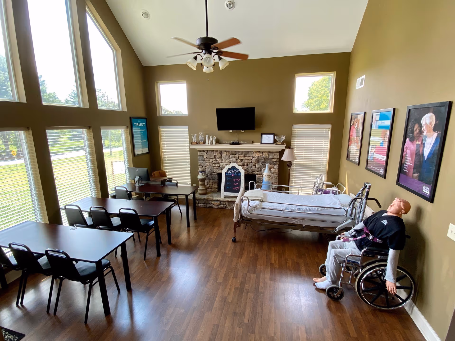 A sunlit senior living room with tables, chairs, a sofa, a hospital bed, and a wheelchair.