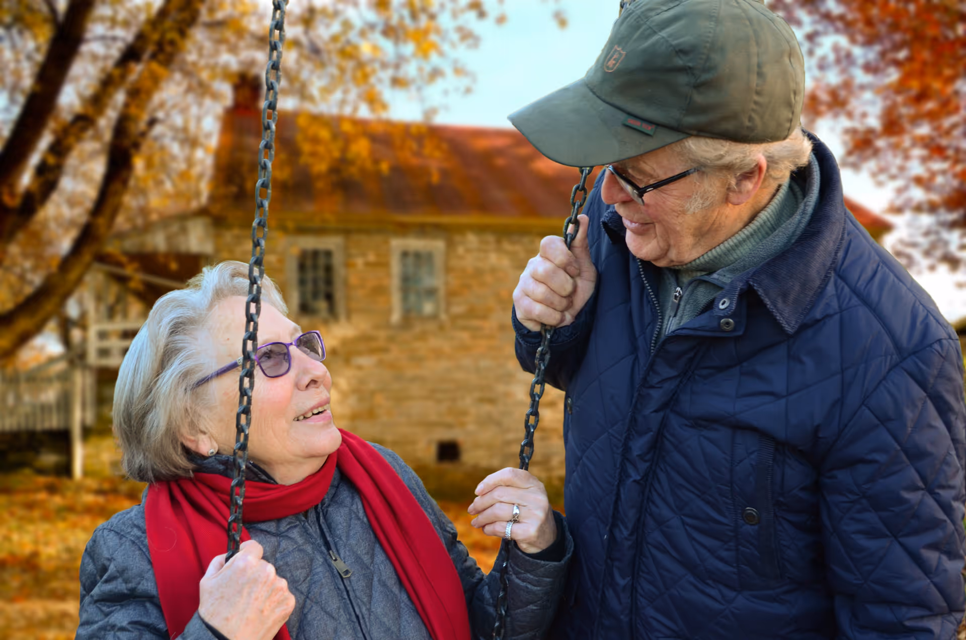 Two elderly people smile on a swing outdoors among autumn trees.