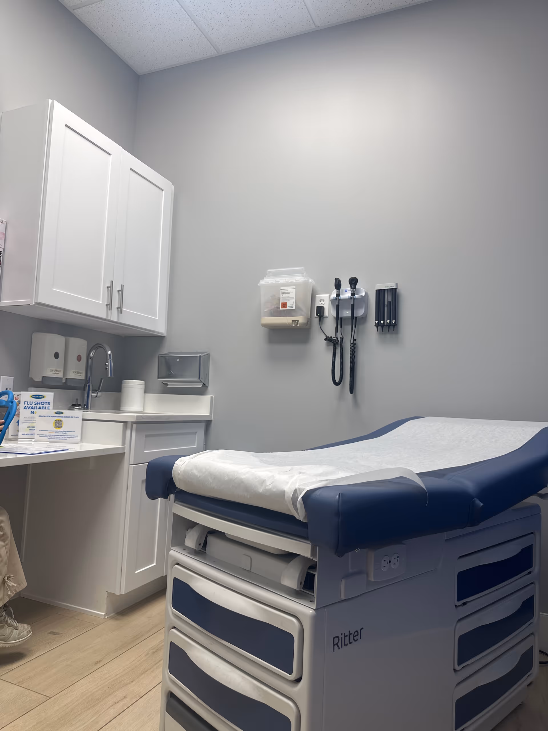 An examination room with a blue patient bed, wall-mounted medical devices, and white cabinetry.