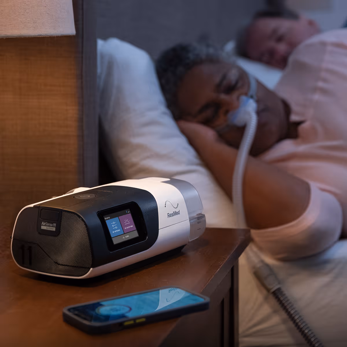 A CPAP machine and smartphone sit on a bedside table beside a sleeping resident.